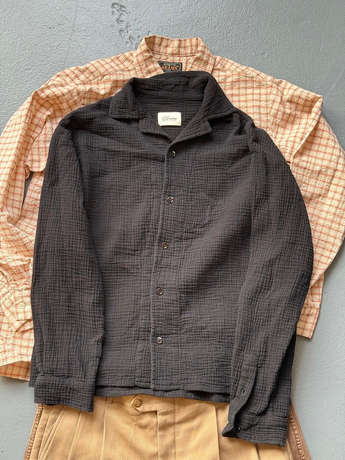 A black textured Ronning shirt laid over a BEAMS check shirt and pleated khaki trousers